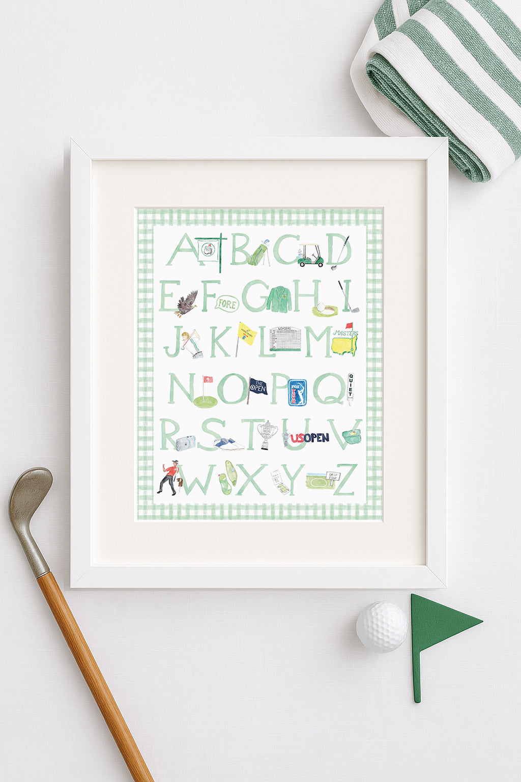 Framed golf alphabet artwork on a white surface with golf accessories.